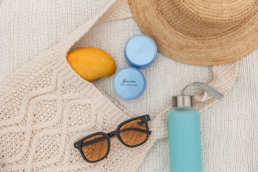 Venice and Miami beach Body butter on the flap of a purse on the beach with a mango, sunglasses, a thermos, and a beach hat.